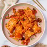 A plate of seared tofu topped with chopped kimchi and sliced green onions, served in a light-colored bowl with chopsticks and a side of rice on a marble surface.