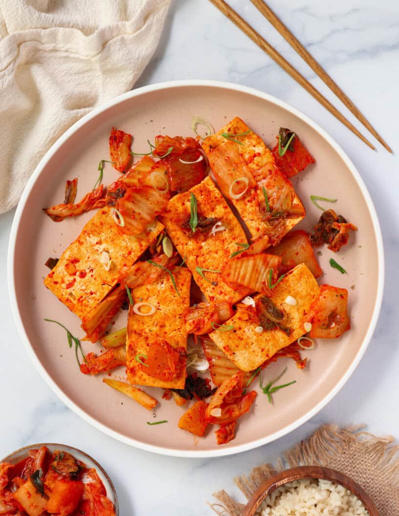 A plate of seared tofu topped with chopped kimchi and sliced green onions, served in a light-colored bowl with chopsticks and a side of rice on a marble surface.