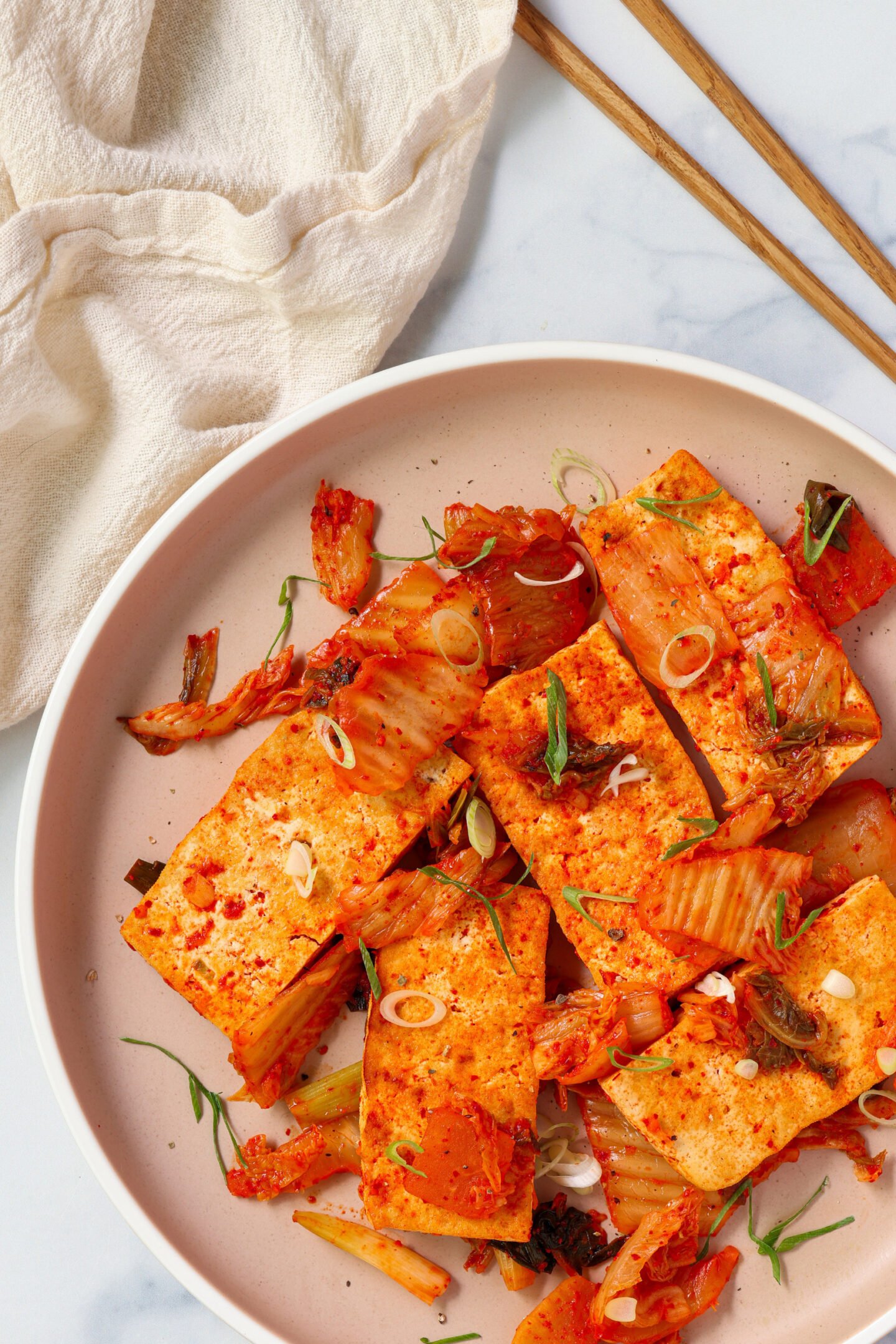 A clean, flat-lay shot of seared tofu and kimchi in a beige plate on a white marble background, garnished with green onions and surrounded by simple props like a napkin and chopsticks.