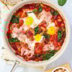 A skillet filled with baked tomatoes, beans, wilted spinach, and perfectly broiled eggs with slices of smoked ham.