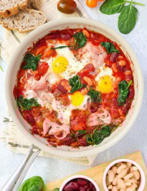 A skillet filled with baked tomatoes, beans, wilted spinach, and perfectly broiled eggs with slices of smoked ham.