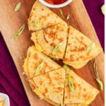 Breakfast quesadillas on a wooden board with sprinkles of green onions, a small cup of salsa, lime wedges and some cheese.