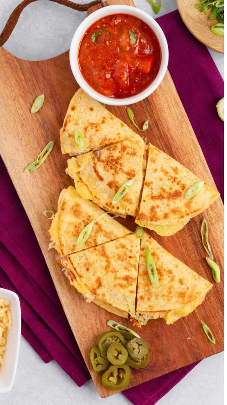 Breakfast quesadillas on a wooden board with sprinkles of green onions, a small cup of salsa, lime wedges and some cheese.