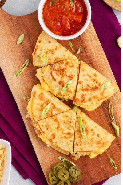 Breakfast quesadillas on a wooden board with sprinkles of green onions, a small cup of salsa, lime wedges and some cheese.