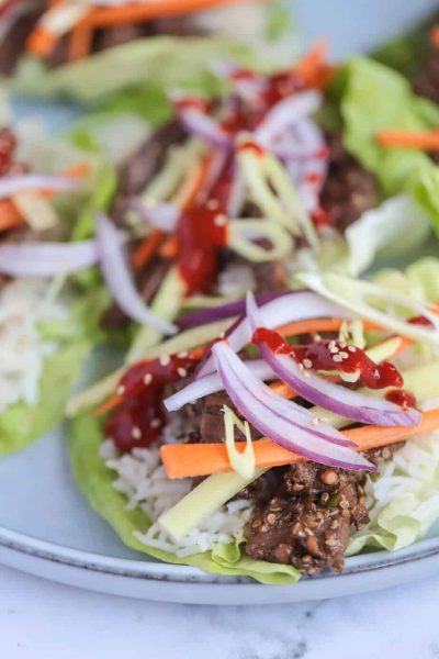 Close-up view of a colorful Korean beef bulgogi lettuce wrap, featuring thinly sliced marinated beef topped with vibrant matchstick-cut carrots, cucumber, thinly sliced red onions, sesame seeds, and a drizzle of gochujang sauce, all nestled in a fresh green lettuce leaf.